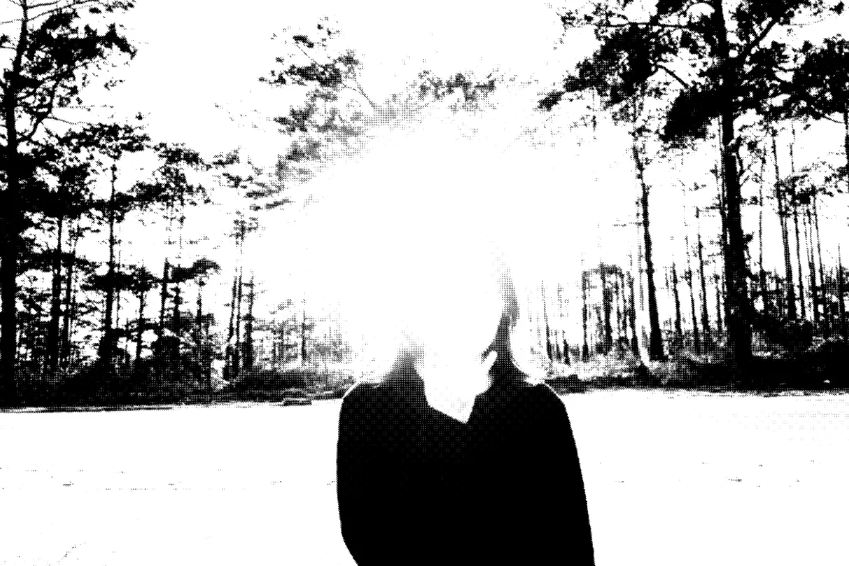 A black-and-white photo of a woman standing in front of the woods, whose face is obstructed by the sunlight.